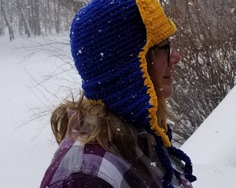 May include: A blue and yellow crocheted hat with ear flaps. The hat is being worn by a person standing in the snow.