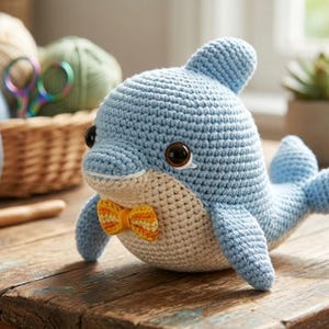 Może przedstawiać: Szydełkowy niebieski delfin z kremowym brzuchem i małą pomarańczową muszką. Ręcznie robiona pluszowa zabawka ma duże, ciemne oczy i jest wystawiona na drewnianej powierzchni. Kosz z przędzą i narzędziami do szydełkowania znajduje się w tle.