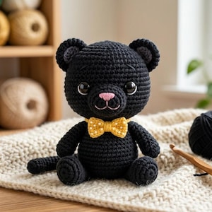 Może przedstawiać: Ręcznie robiony czarny szydełkowy miś z żółtą muszką i różowym nosem siedzi na kremowym, teksturowanym kocu. W pobliżu znajduje się kulka czarnej przędzy i szydełko. Tło obejmuje przędzę i roślinę sukulent.