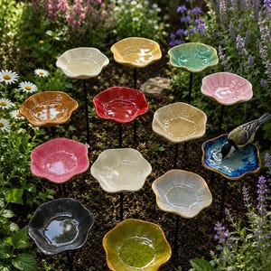 May include: A collection of twelve ceramic flower-shaped bird baths in various colors including yellow, red, blue, pink, green, and grey. Each bowl is mounted on a thin black metal stake, arranged in a garden setting with a small bird drinking.