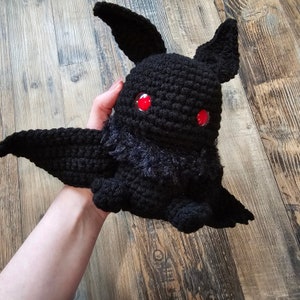 May include: A handmade black crochet moth plushie with red eyes and feathery chest detail. The plushie has large wings and pointed ears. The moth is held in a person's hand against a wooden background.