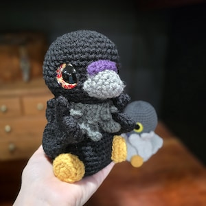 May include: A black crocheted bird plush with a gray beak, a purple patch above the beak, and yellow feet. The bird has large, glossy eyes with a red and orange patterned iris. A smaller, gray and white crocheted bird plush sits in the background.