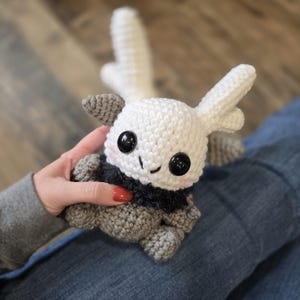 May include: A handmade crochet plush toy in the shape of a creature with large black eyes, small gray ears, and white wings. The toy has a white head and wings, a gray body, and a black collar. The toy is being held.
