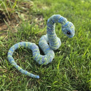 Peut inclure: Un jouet serpent crocheté bleu et vert, posé sur de l'herbe verte. Le serpent a un corps courbé et une petite tête ronde avec un œil sombre. Le fil a un aspect texturé.