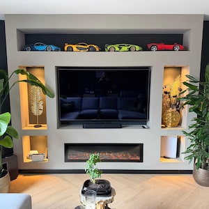 Op de afbeelding: Een moderne woonkamer met een ingebouwd entertainmentcenter. Het center heeft een grote flatscreen-tv, een open haard en inbouwkasten. Vier speelgoed sportwagens in blauw, geel, groen en rood staan op een bovenste plank. Potplanten voegen groen toe.