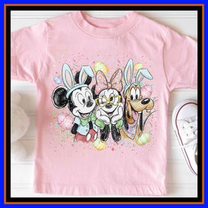 May include: Pink toddler t-shirt featuring Mickey Mouse, Minnie Mouse, and Goofy dressed as Easter bunnies. The graphic includes colourful Easter eggs and confetti. A white trainer is visible on the right side of the image.
