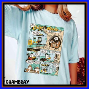 Könnte beinhalten: Hellblaues T-Shirt mit einem Comic-Strip mit Chip und Chap-Charakteren. Der Comic-Strip zeigt Donald Duck und die Chipmunks in einer Campingszene. Das Shirt hat den Text "Chip 'n Dale The Rescue Rangers" und das Wort "CHAMBRAY" unten.