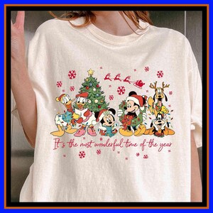 Może przedstawiać: Kremowa koszulka z świątecznym motywem z postaciami Disneya. Grafika zawiera Myszkę Miki i Minnie, Kaczora Donalda i Daisy, Goofy'ego i Pluto, choinkę, płatki śniegu i napis "It's the most wonderful time of the year."