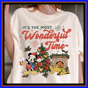 Może przedstawiać: Kremowa koszulka z napisem "It's the Most Wonderful Time". Grafika przedstawia Myszka Miki w czapce Świętego Mikołaja, choinkę, kominek i Psa Pluto. Projekt jest w kolorach czerwonym, zielonym i żółtym, idealny na święta.