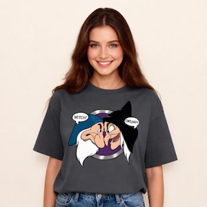 Könnte beinhalten: Dunkelgraues T-Shirt mit einem Cartoon-Motiv von zwei Figuren mit den Textblasen "WITCH!" und "GRUMPY!". Die Grafik befindet sich in einem silbernen Kreis. Das Shirt hat einen Rundhalsausschnitt und kurze Ärmel.