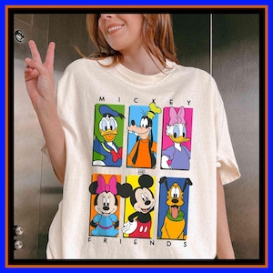 Może przedstawiać: Kremowa koszulka z kolorową grafiką Myszki Miki i przyjaciół. Projekt zawiera Kaczora Donalda, Goofy'ego, Daisy, Minnie, Myszka Miki i Pluto. Napis "MICKEY AND FRIENDS" jest wydrukowany nad i pod postaciami.