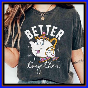 Puede incluir: Camiseta gris oscuro con un diseño de tetera y taza de dibujos animados, y la frase blanca "BETTER together". La tetera y la taza tienen caras sonrientes y están delineadas en negro, con detalles dorados, rosas y morados. Una camiseta de estilo casual.