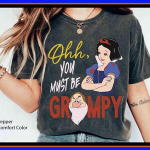 Peut inclure: T-shirt Comfort Color poivre avec un motif de Blanche-Neige et Grincheux et le texte "Ohh, you must be grumpy". Le design comprend des couleurs jaune, rouge, bleu et blanc.