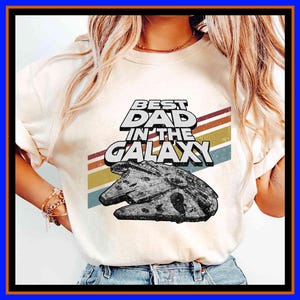 Può includere: T-shirt color crema con la scritta "BEST DAD IN THE GALAXY" in un font retrò, con una grafica di un'astronave. Il design include uno sfondo a strisce nei toni del marrone, giallo e blu. Una maglietta casual e confortevole.