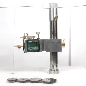 Pu&ograve; includere: Un calibro di altezza in metallo argentato dotato di display digitale con accenti verdi, manopole di regolazione in ottone e una colonna verticale. Quattro dischi metallici circolari simili a ingranaggi con bordi dentellati sono allineati.