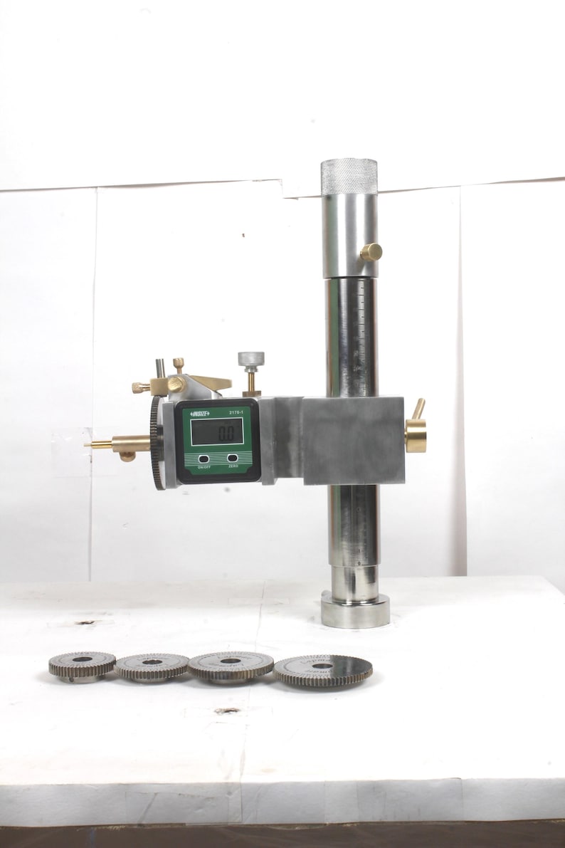 Pu&ograve; includere: Un calibro di altezza in metallo argentato dotato di display digitale con accenti verdi, manopole di regolazione in ottone e una colonna verticale. Quattro dischi metallici circolari simili a ingranaggi con bordi dentellati sono allineati.