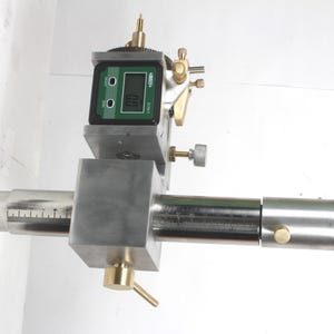 Pu&ograve; includere: Uno strumento di misurazione di precisione in metallo argentato dotato di un display digitale con sfondo verde e testo nero che indica 0.0. Il dispositivo include una barra cilindrica orizzontale con segni di misurazione incisi, manopole di regolazione in ottone e un'estremit&agrave; zigrinata.