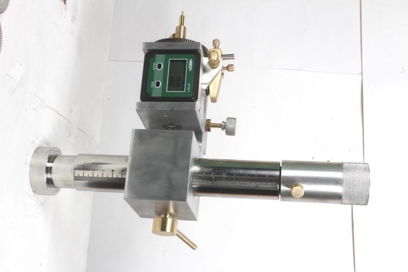 Pu&ograve; includere: Uno strumento di misurazione di precisione in metallo argentato dotato di un display digitale con sfondo verde e testo nero che indica 0.0. Il dispositivo include una barra cilindrica orizzontale con segni di misurazione incisi, manopole di regolazione in ottone e un'estremit&agrave; zigrinata.