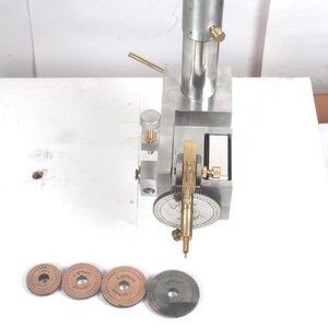 Pu&ograve; includere: Un utensile di precisione in metallo argentato con un montante cilindrico verticale, un meccanismo a ingranaggi centrale e manopole di regolazione in ottone. Quattro dischi metallici circolari dentati marcati Lapmart sono in fila.