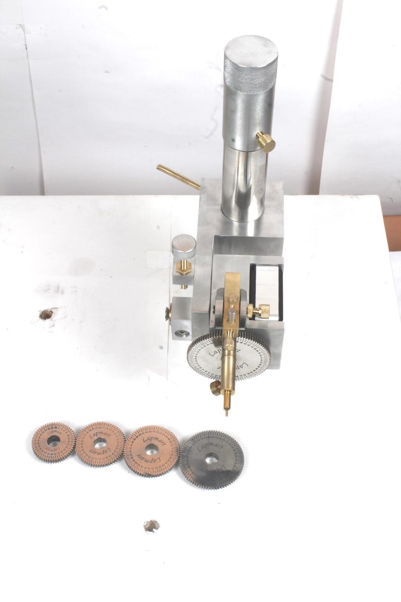 Pu&ograve; includere: Un utensile di precisione in metallo argentato con un montante cilindrico verticale, un meccanismo a ingranaggi centrale e manopole di regolazione in ottone. Quattro dischi metallici circolari dentati marcati Lapmart sono in fila.