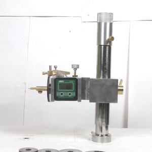Pu&ograve; includere: Un calibro di altezza in metallo con un display digitale che mostra 0.0, una colonna cilindrica verticale e manopole di regolazione in ottone. Quattro ingranaggi metallici circolari dentati sono disposti in fila davanti al dispositivo.