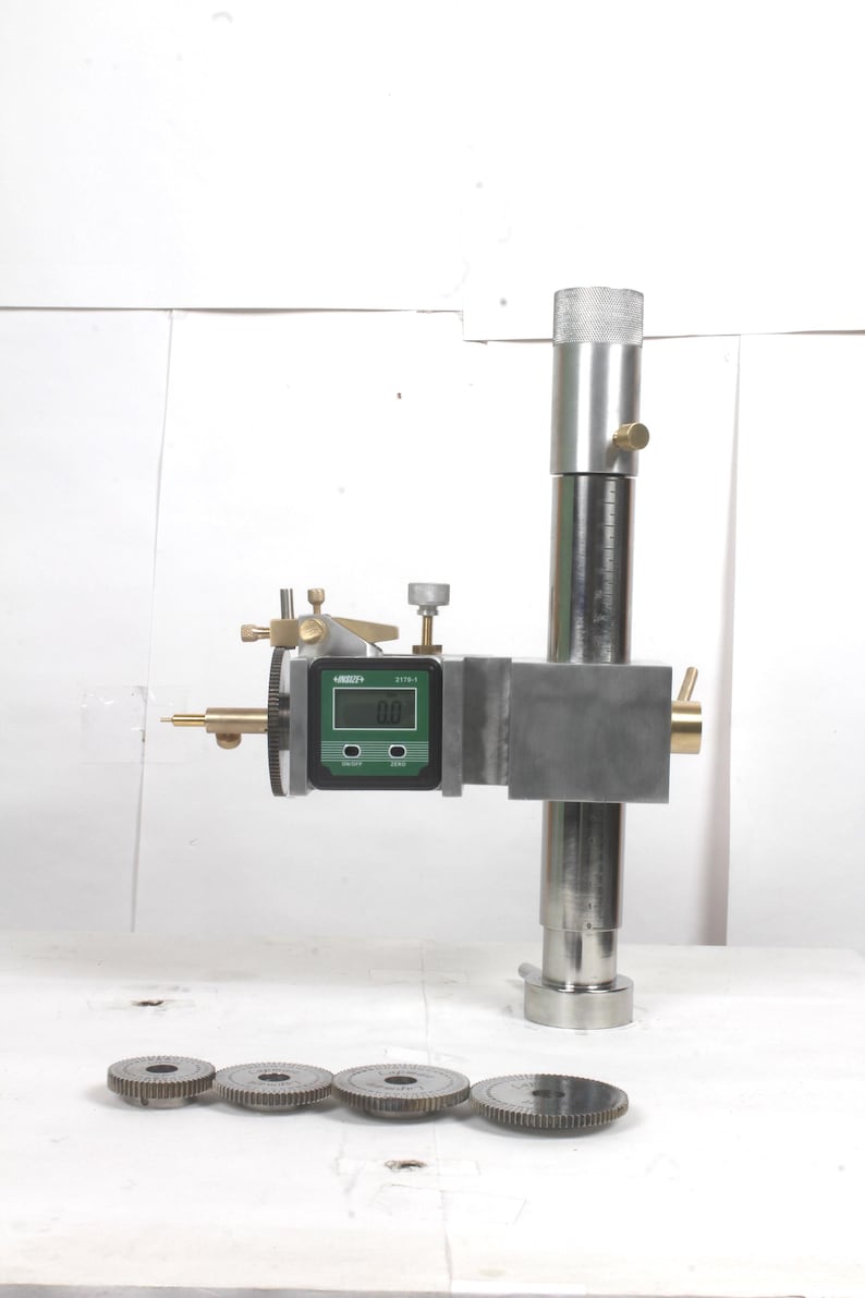 Pu&ograve; includere: Un calibro di altezza in metallo con un display digitale che mostra 0.0, una colonna cilindrica verticale e manopole di regolazione in ottone. Quattro ingranaggi metallici circolari dentati sono disposti in fila davanti al dispositivo.