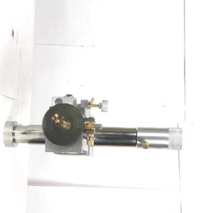 Pu&ograve; includere: Un accessorio per tornio in metallo color argento dotato di un albero cilindrico centrale, un ingranaggio circolare scuro con dettagli in ottone e una base quadrata. Quattro ingranaggi metallici circolari di varie dimensioni sono disposti in linea verticale.