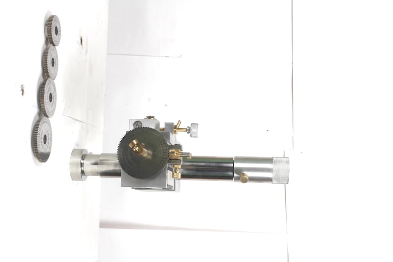 Pu&ograve; includere: Un accessorio per tornio in metallo color argento dotato di un albero cilindrico centrale, un ingranaggio circolare scuro con dettagli in ottone e una base quadrata. Quattro ingranaggi metallici circolari di varie dimensioni sono disposti in linea verticale.