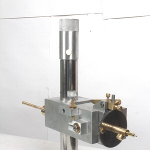 Pu&ograve; includere: Un utensile di precisione in metallo caratterizzato da un montante cilindrico verticale con parte superiore zigrinata, un blocco rettangolare centrale e un gruppo montato lateralmente con ingranaggio nero, manopole in ottone e punta metallica.