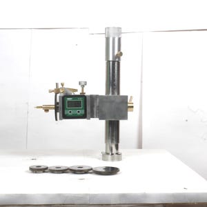 Pu&ograve; includere: Un calibro di altezza in metallo argentato dotato di display digitale con dettagli verdi, una colonna verticale e un braccio orizzontale. Quattro ingranaggi metallici circolari di varie dimensioni sono disposti in fila davanti allo strumento.