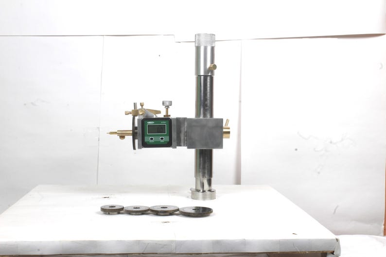 Pu&ograve; includere: Un calibro di altezza in metallo argentato dotato di display digitale con dettagli verdi, una colonna verticale e un braccio orizzontale. Quattro ingranaggi metallici circolari di varie dimensioni sono disposti in fila davanti allo strumento.