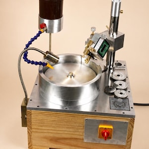 Peut inclure: Une machine à facetter les pierres précieuses avec une base en bois, un plateau métallique circulaire, une jauge d'angle numérique et un tuyau de refroidissement bleu flexible. L'unité comprend un mât vertical, un cadran de vitesse et un interrupteur rouge.