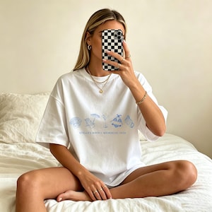 May include: White t-shirt with a blue beach-themed graphic and the text "STELLA'S BACH / WAIKIKI HI 2024". The shirt features illustrations of seashells, beach chairs, and swimwear. The model is wearing gold jewelry.