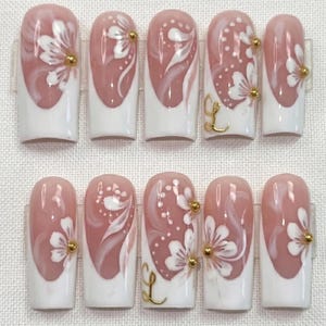May include: A set of ten artificial nails. Each nail has a white base and a pinkish-nude top, decorated with white floral patterns and gold accents. Some nails feature a gold letter "L".