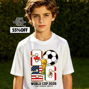 May include: A white t-shirt featuring the text "2026" with a soccer ball graphic and a golden trophy. The design incorporates the flags of Canada, the United States, and Mexico, with the words "World Cup 2026" and "USA CANADA MEXICO" below.