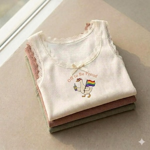 Puede incluir: Una pila de tres camisetas sin mangas en crema, malva y verde oliva. La camiseta superior de color crema presenta un ribete de encaje, un pequeño lazo y un ganso bordado que sostiene una bebida y una bandera arcoíris, con el texto "Off to Be Proud".