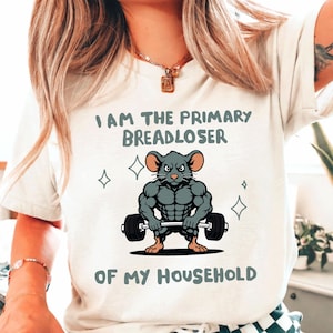 May include: Off-white t-shirt with the text "I AM THE PRIMARY BREADLOSER OF MY HOUSEHOLD." A muscular gray mouse graphic is lifting a barbell. The design includes four diamond shapes.