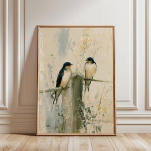 Op de afbeelding: Ingelijste kunstdruk met twee zwaluwen op een verweerde houten hek. De vogels hebben een zwart-witte verenkleed, tegen een zachte, gedempte achtergrond met bloemaccenten. Het kunstwerk zit in een houten lijst.