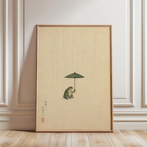 Peut inclure: Impression d'art encadrée représentant une grenouille verte tenant un petit parapluie vert sous la pluie. L'œuvre a une esthétique vintage avec du texte japonais et un sceau rouge. Le fond est beige clair.