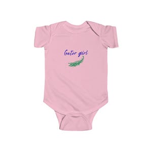 May include: A light pink baby bodysuit with short sleeves. The text "Gator girl" is written in a cursive font in blue, with a green alligator illustration below the text. The bodysuit is made of soft material.