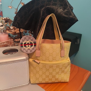 May include: A yellow handbag with a matching coin purse. The handbag features a repeating logo pattern and pink accents. The coin purse is round with a logo design and red and green stripes. A black bag is in the background.