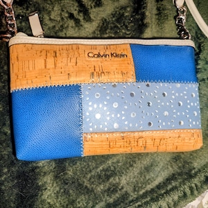 Könnte beinhalten: Eine Patchwork-Umhängetasche mit blauen, Kork- und hellblauen Abschnitten. Die Tasche hat einen silbernen Kettenriemen und einen Reißverschluss. Das "Calvin Klein"-Logo ist sichtbar. Das Design kombiniert Materialien und Farben.