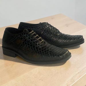 May include: A pair of black lace-up shoes featuring a textured, scale-like pattern across the entire upper surface. The shoes have a low-profile silhouette, a rounded toe box, and dark laces that match the deep black tone of the exterior material.
