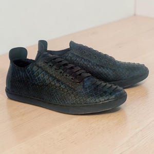 Puede incluir: Un par de zapatillas bajas de color azul oscuro, con un patrón texturizado similar a escamas en la parte superior. El calzado incluye cordones negros, punta redondeada y una suela plana de goma azul oscuro a juego con una lengüeta trasera.