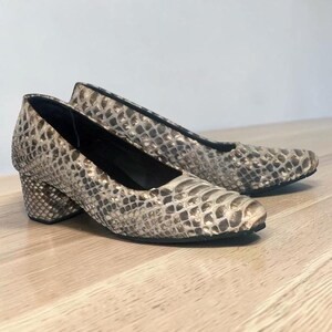 May include: A pair of low-heeled pumps featuring a textured snakeskin pattern in shades of beige, brown, and cream. The shoes have a rounded toe, a block heel, and a black interior lining, showcasing a consistent reptilian scale design throughout.