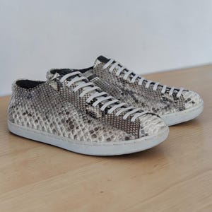 May include: A pair of low-top sneakers featuring a grey and white snakeskin-patterned exterior. The shoes have a rounded toe, white laces threaded through eyelets, and a solid white rubber sole that provides a clean contrast to the textured upper.