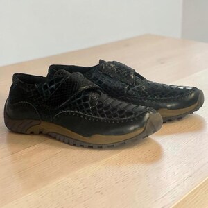 May include: A pair of black shoes featuring a textured snakeskin-like pattern on the upper, a hook-and-loop closure strap across the top, and a brown rubber sole with a treaded design. The shoes have a low-top silhouette and rounded toes.