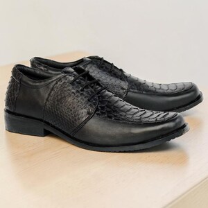 May include: A pair of black lace-up dress shoes featuring a combination of smooth leather and textured snakeskin-patterned panels. The shoes have a rounded toe, a low heel, and black laces, showcasing a sleek design with distinct material contrast.