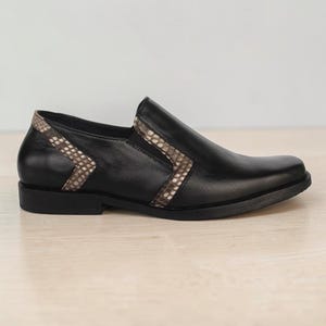 Peut inclure: Une chaussure à enfiler noire avec une finition lisse et un panneau en forme de V distinctif en matériau texturé à motif peau de serpent dans des tons marron et beige sur le côté et le col, posée sur une semelle noire avec un petit talon plat.