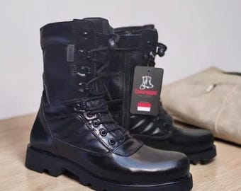 May include: A pair of black leather combat boots with laces and a side zipper. The boots have a thick sole and a tag with a logo. The boots are on a wooden surface.